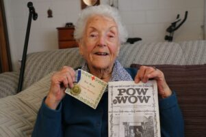 Mildred Turley holding one of her husband Trevor’s service medals, a card that was attached to a parcel sent by the British Red Cross to POWs, and the volume of the POW WOW journal which contained Trevor’s obituary.